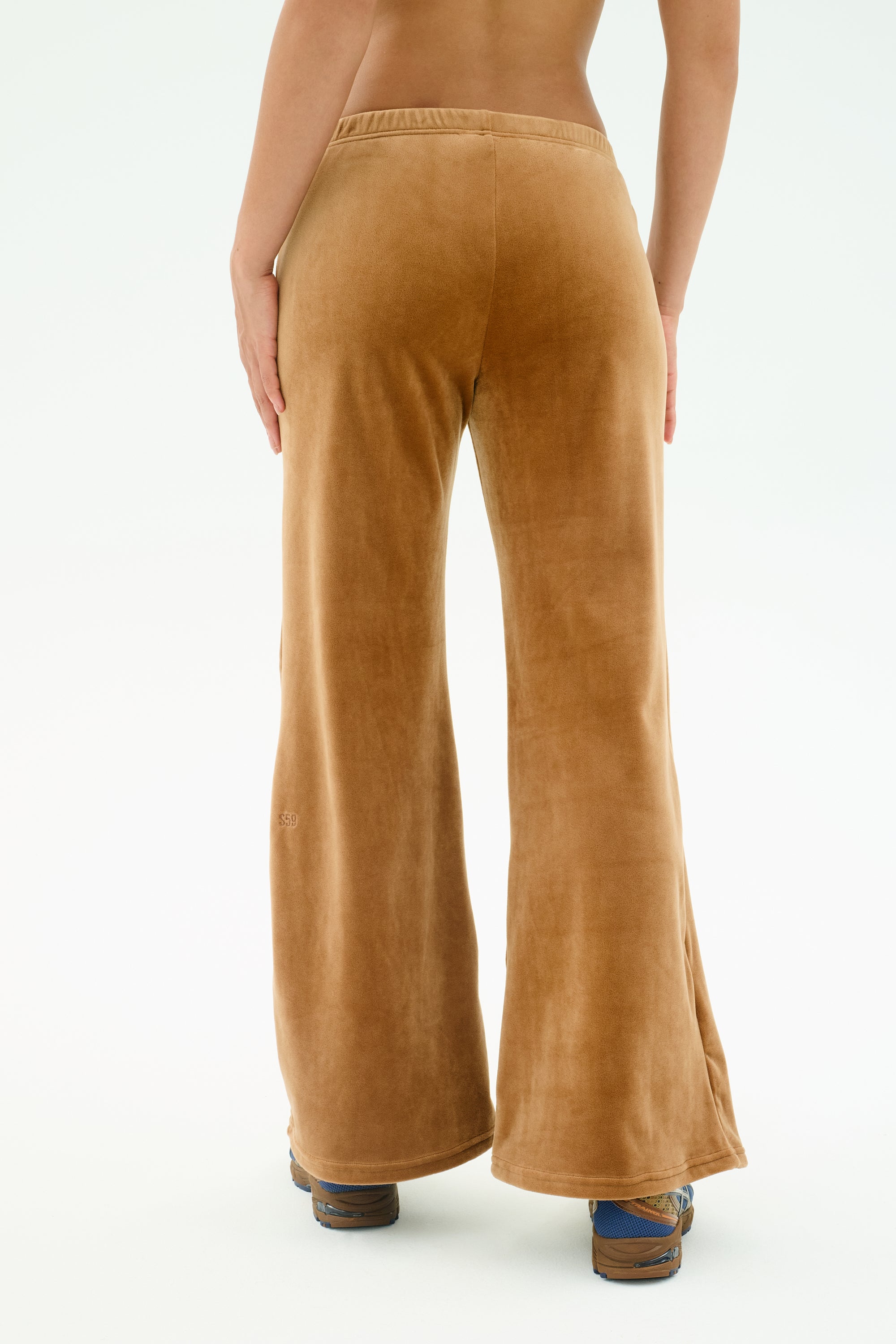 A person stands facing away from the camera, wearing SPLITS59's Joy Velour Pant in Honey—high-waisted, wide-leg brown pants made of polyester-spandex velour—paired with patterned shoes against a plain white background.