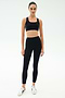 A person stands facing forward, wearing a black sports bra, SPLITS59 Margot Rigor Legging - Black/Dark Chocolate, and white sneakers against a plain light background.