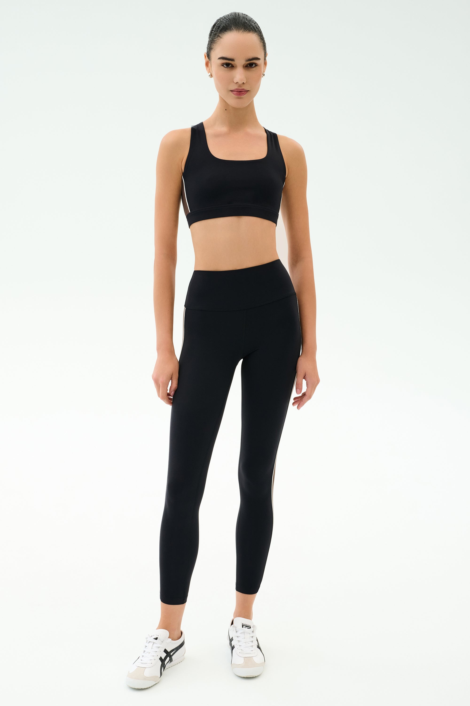 A person stands facing forward, wearing a black sports bra, SPLITS59 Margot Rigor Legging - Black/Dark Chocolate, and white sneakers against a plain light background.