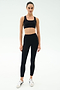 A person stands facing forward, wearing a black sports bra, SPLITS59 Margot Rigor Legging - Black/Dark Chocolate, and white sneakers against a plain light background.