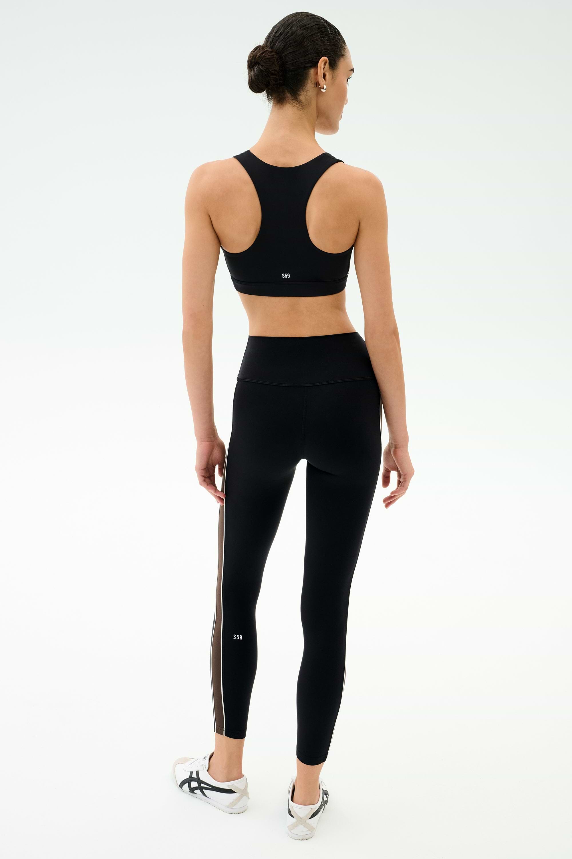 A woman faces away wearing a black sports bra, SPLITS59 Margot Rigor Legging in Black/Dark Chocolate, and white athletic shoes against a plain light background.