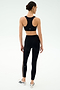 A woman faces away wearing a black sports bra, SPLITS59 Margot Rigor Legging in Black/Dark Chocolate, and white athletic shoes against a plain light background.