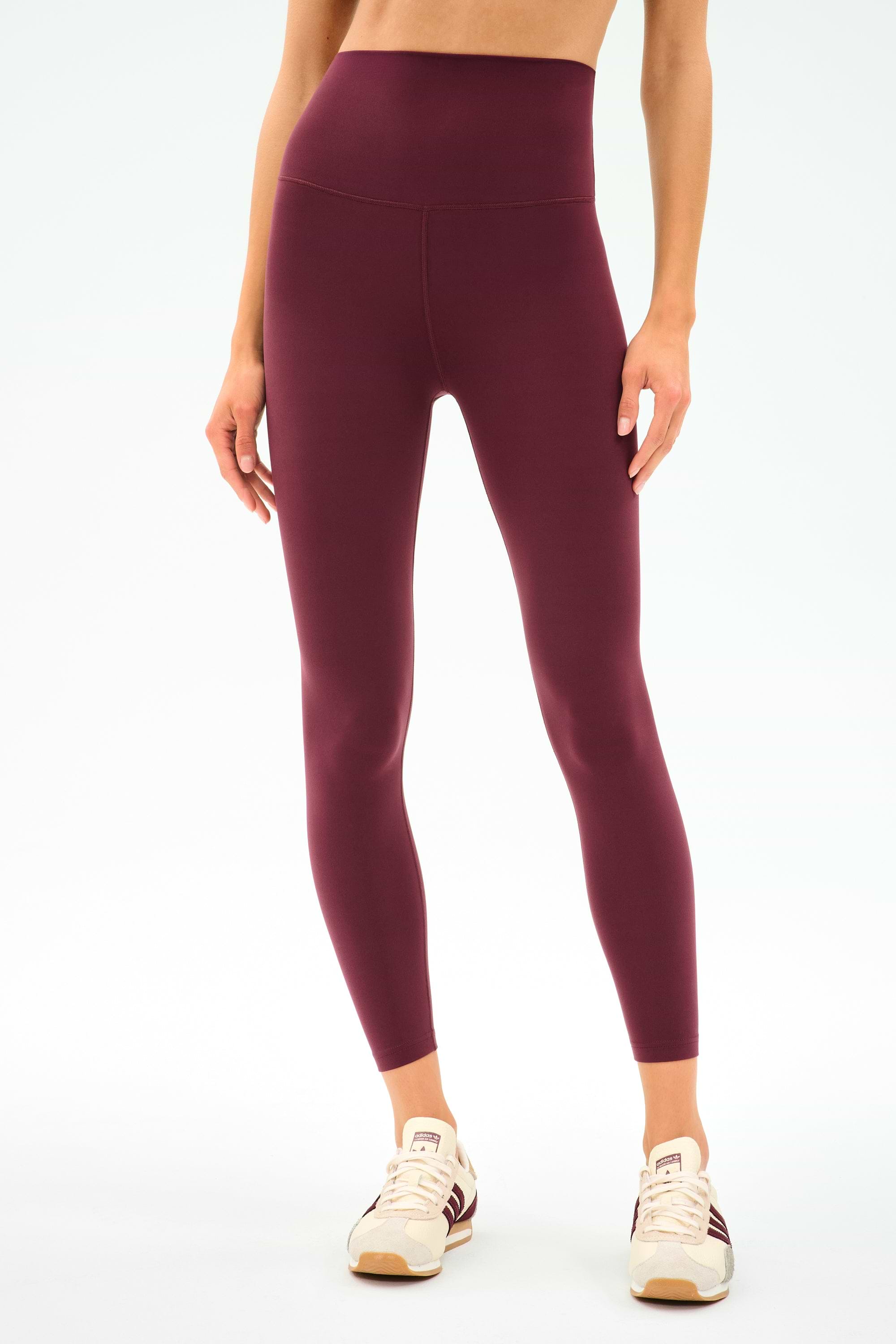 A person wears SPLITS59 Airweight Super High Waist 7/8 leggings in Dark Cherry and cream sneakers against a white background—ideal for hot yoga or everyday comfort.