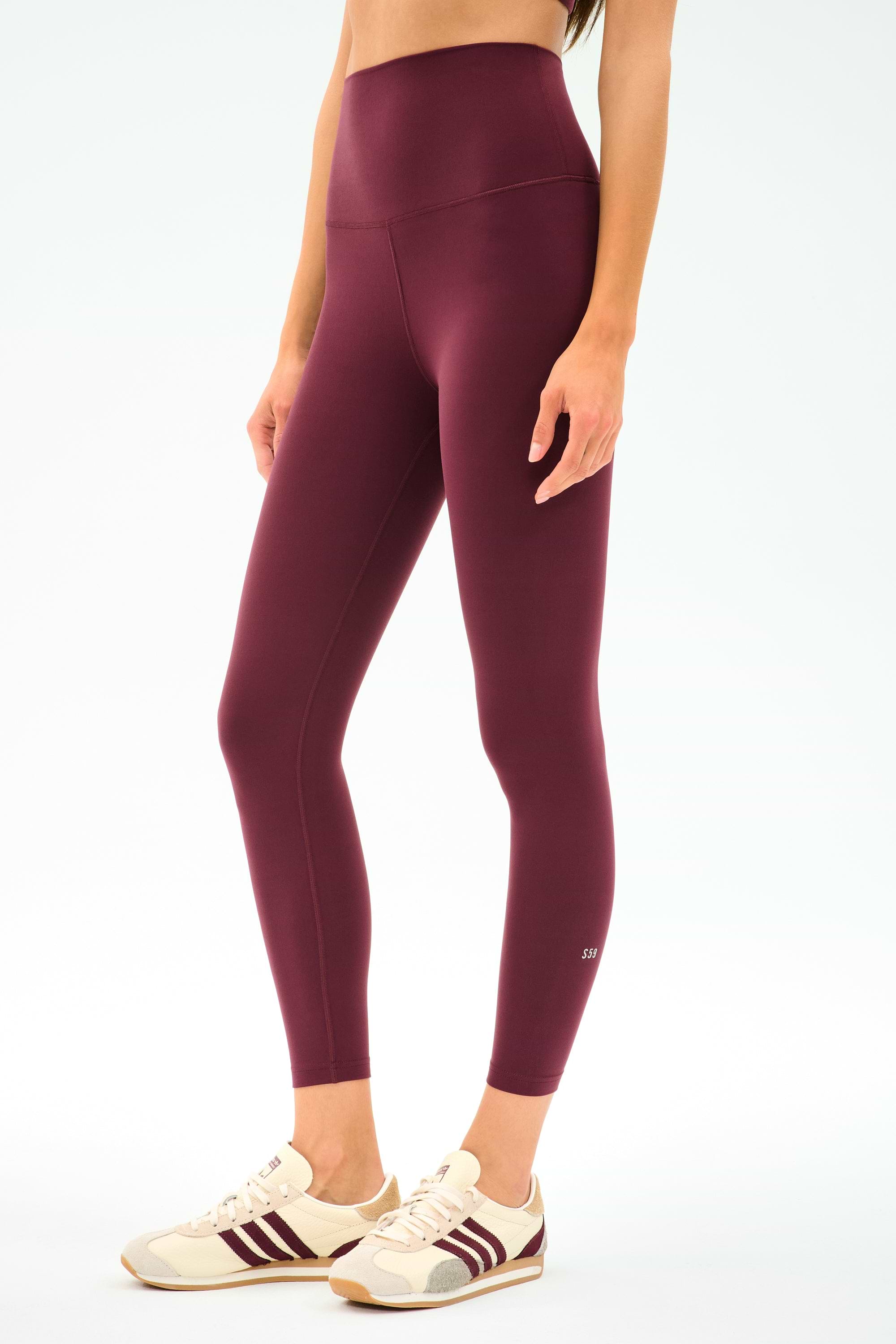 A woman wears SPLITS59 Airweight Super High Waist 7/8 leggings in Dark Cherry with cream sneakers featuring maroon stripes, standing against a plain white background.