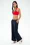 A woman stands in a studio wearing the SPLITS59 Michele Airweight Trouser in Indigo, paired with a red sports bra and white sneakers with red stripes, her hands tucked into her pockets.