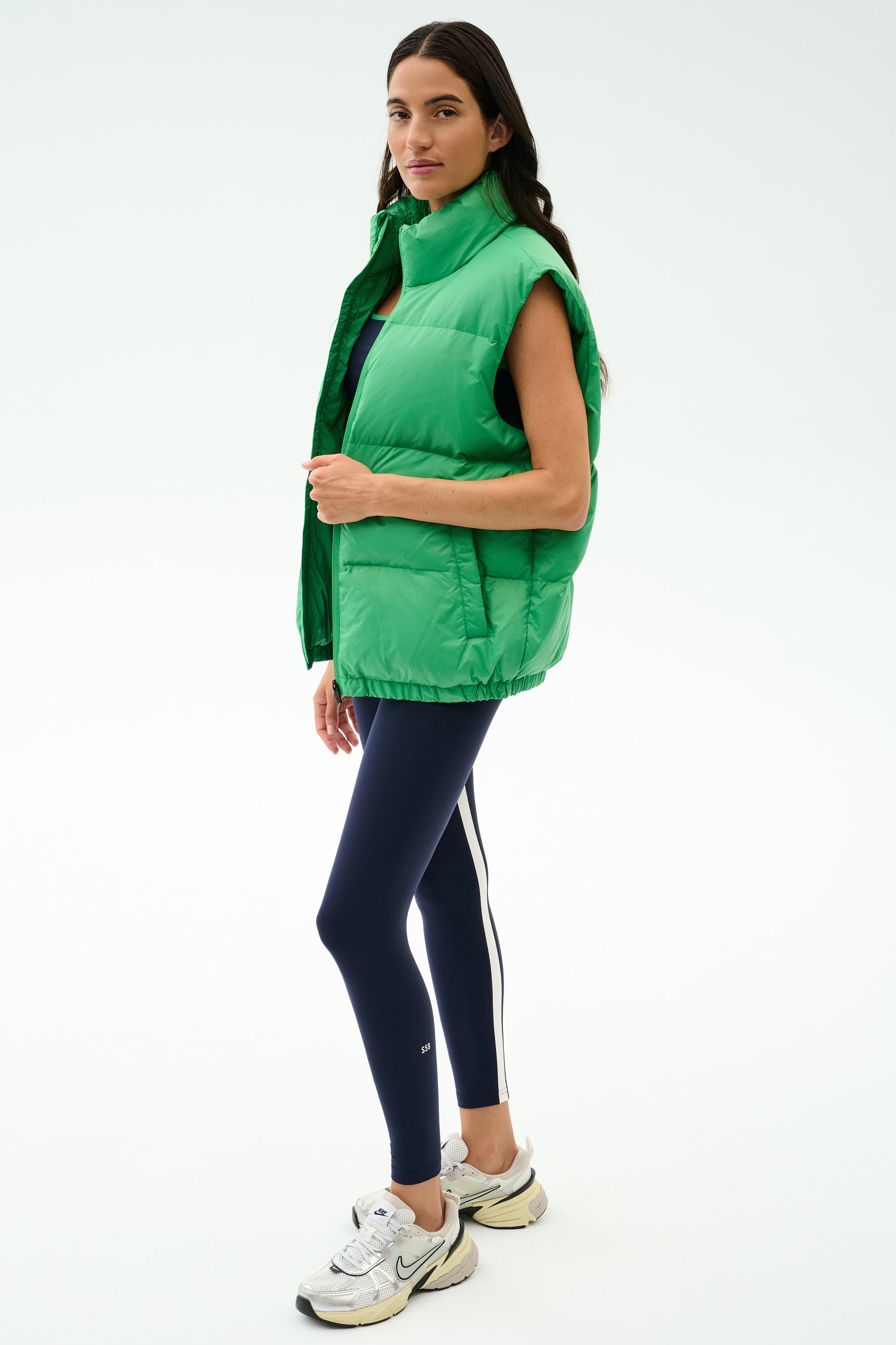 A woman wears the SPLITS59 Summit Nylon Puffer Vest in Grass for lightweight warmth, paired with navy leggings featuring a white stripe and white sneakers—ideal for cool weather workouts against a plain light backdrop.