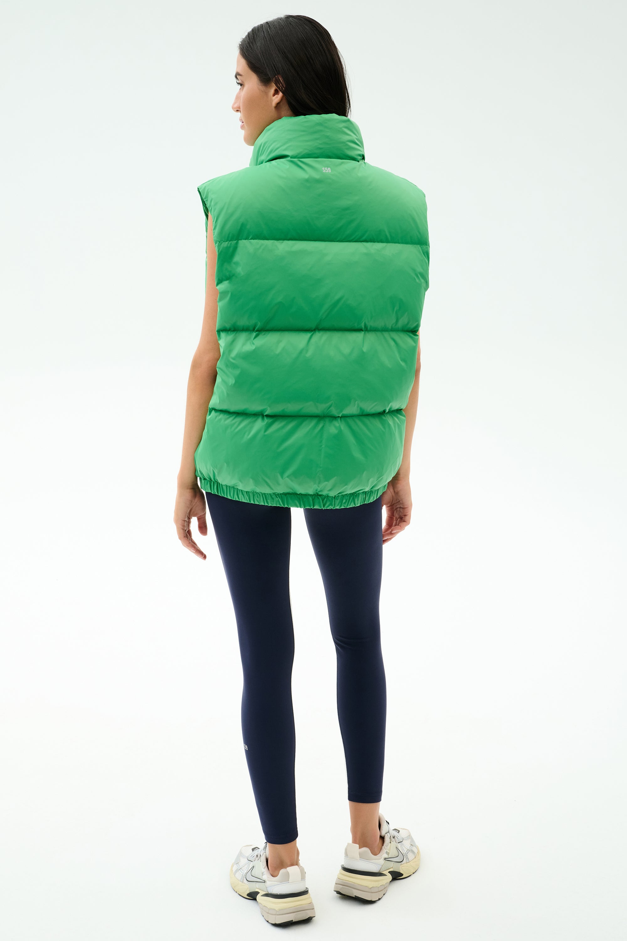 A woman faces away, wearing the SPLITS59 Summit Nylon Puffer Vest in Grass—a sleeveless, lightweight insulated vest—paired with dark leggings and sneakers, set against a plain light background.