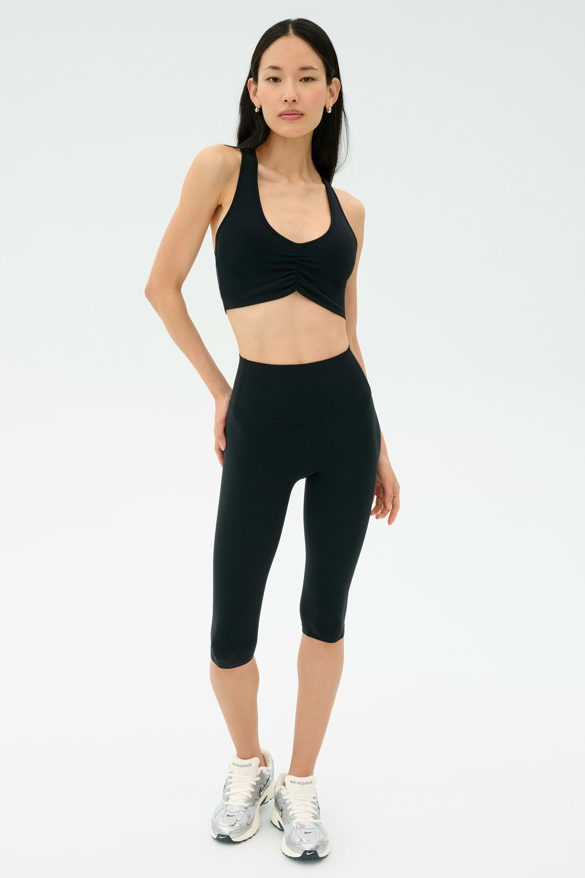 A woman stands against a plain background wearing a black sports bra, SPLITS59 Airweight High Waist 16" Legging in black, and white athletic shoes.