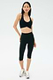 A person stands against a plain background wearing a black SPLITS59 Airweight Bra with Ruching, black high-waisted capri leggings, and gray running shoes.