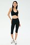 A person stands against a plain background wearing a black SPLITS59 Airweight Bra with Ruching, black high-waisted capri leggings, and gray running shoes.