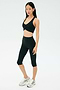 A woman stands against a plain background wearing the SPLITS59 Airweight Bra with Ruching in black, paired with black capri leggings and silver athletic shoes.