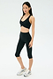 A woman stands against a plain background wearing the SPLITS59 Airweight Bra with Ruching in black, paired with black capri leggings and silver athletic shoes.