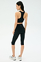 A woman faces away, wearing the SPLITS59 Airweight Bra with Ruching in Black, paired with black capri leggings and silver sneakers against a plain light background.