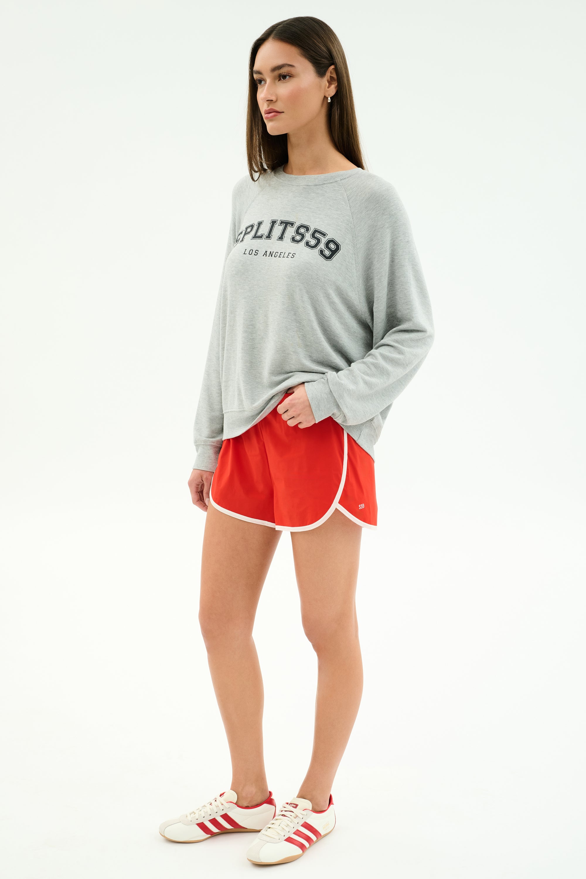 A woman wears the SPLITS59 Andie Oversized Fleece Varsity Graphic Sweatshirt in Heather Grey and red athletic shorts, styled with white sneakers. This cozy Los Angeles-made look features comfort and varsity flair.