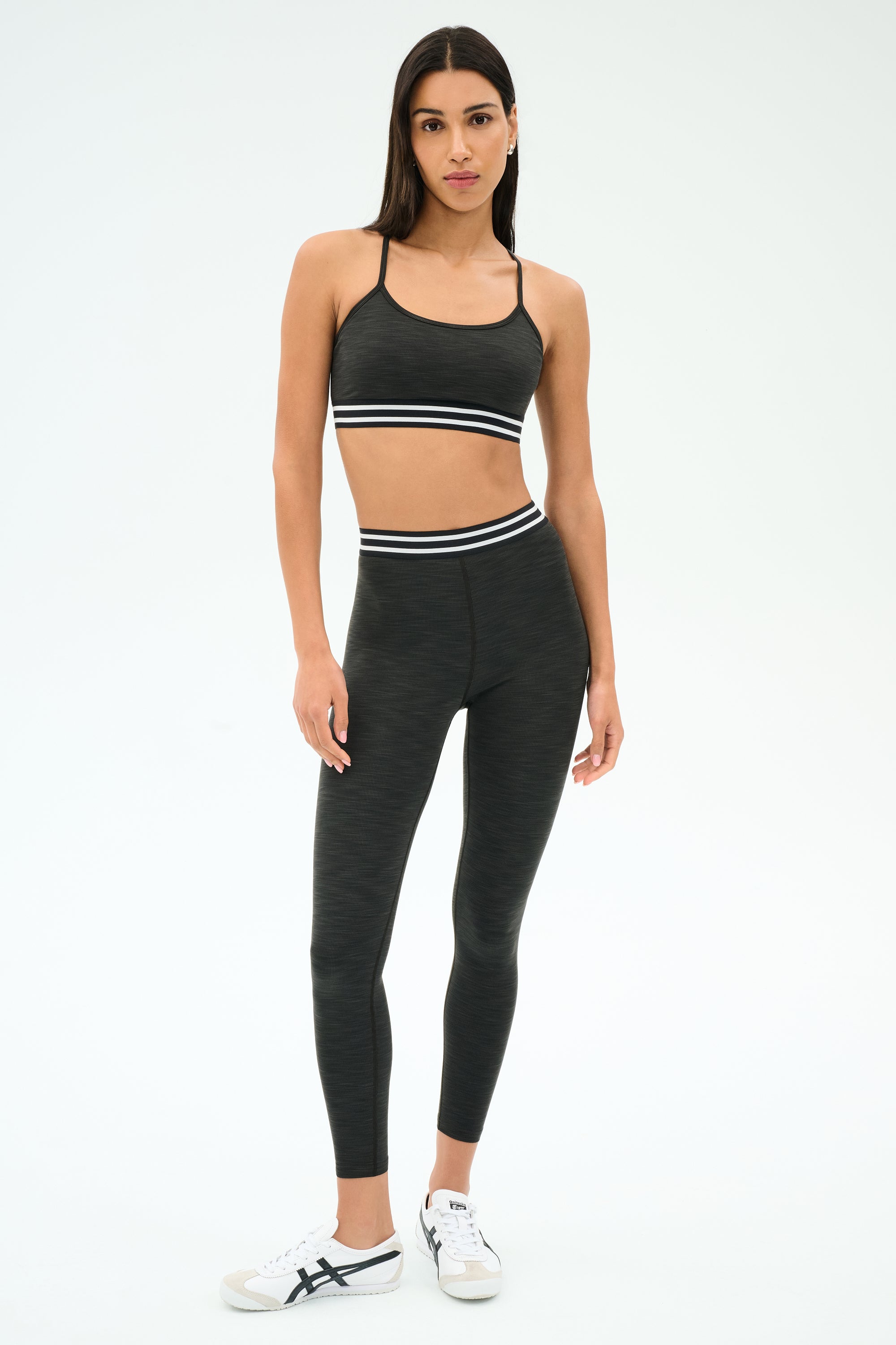 A woman stands against a plain background wearing the SPLITS59 Cali Active Rib Bra in Heather Graphite, paired with matching leggings featuring white trim and white sneakers.