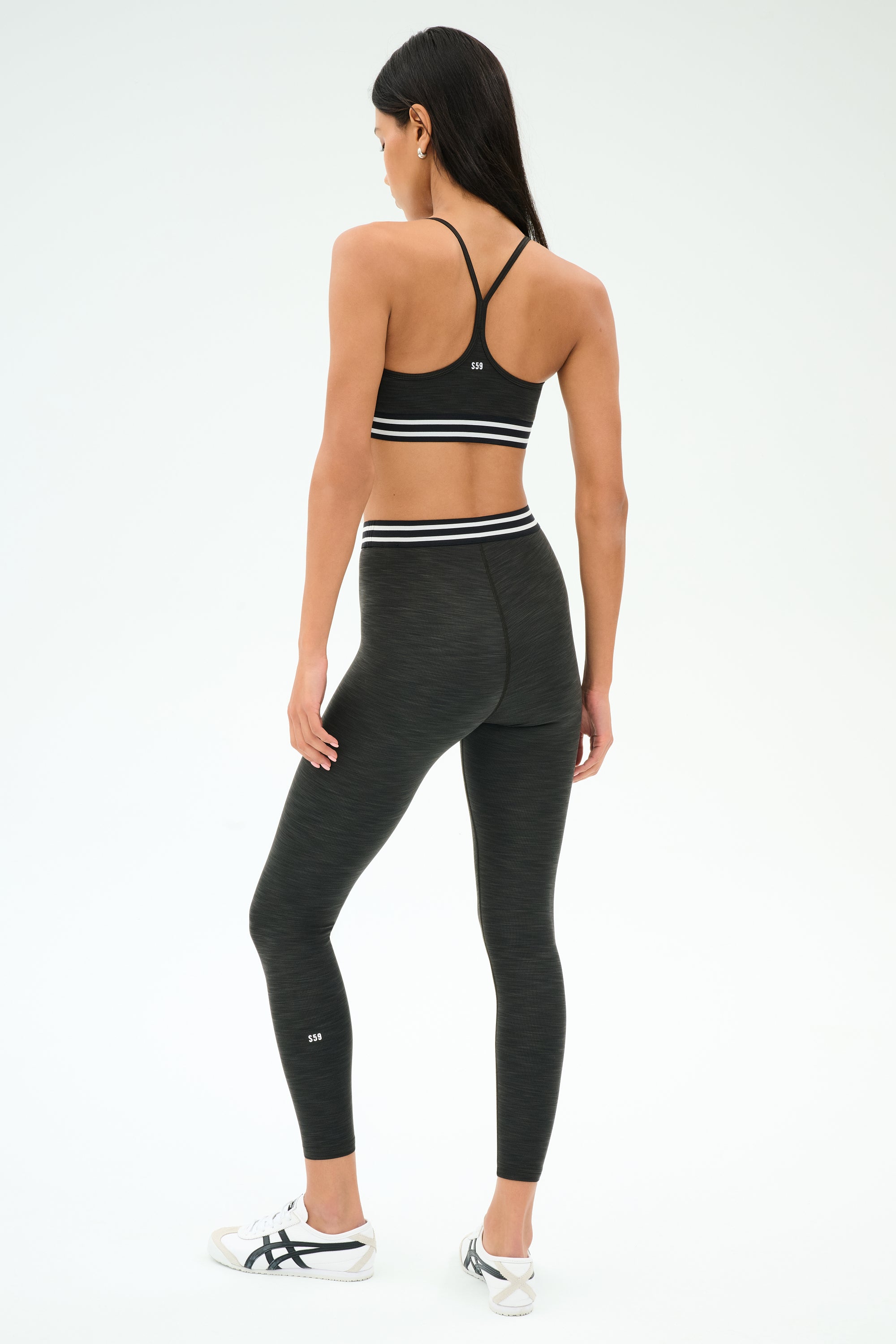 A woman faces away, wearing the SPLITS59 Cali Active Rib Bra in Heather Graphite, paired with black high-waisted leggings and white sneakers against a plain white background.