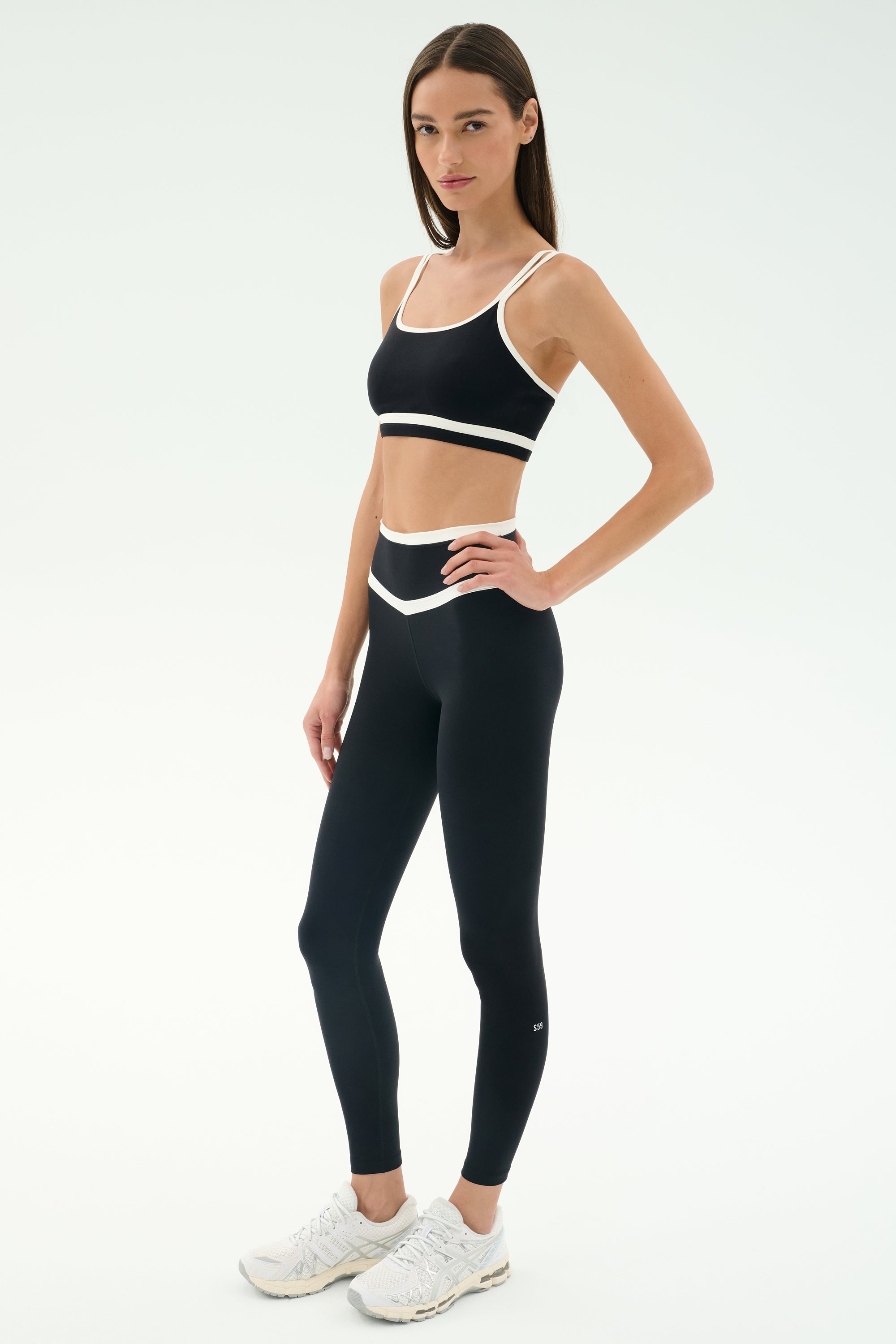 A woman wears the SPLITS59 Mason High Waist Rigor Full Length leggings in Black/White, paired with a black sports bra and white sneakers, standing against a plain light background.