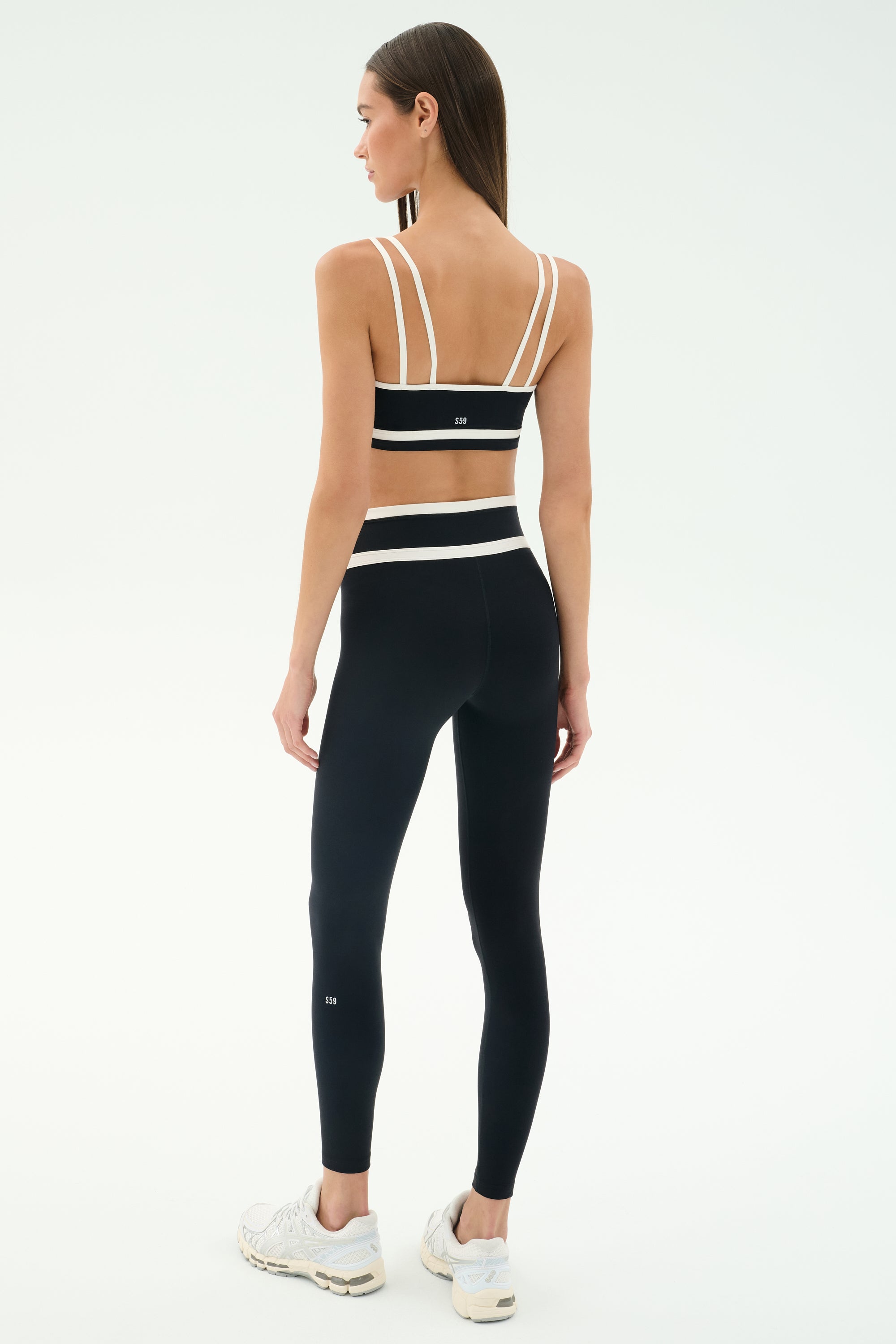 A woman stands facing away, ready to work out in SPLITS59's Mason High Waist Rigor Full Length leggings in Black/White and a black sports bra with white straps, paired with white sneakers against a plain background.
