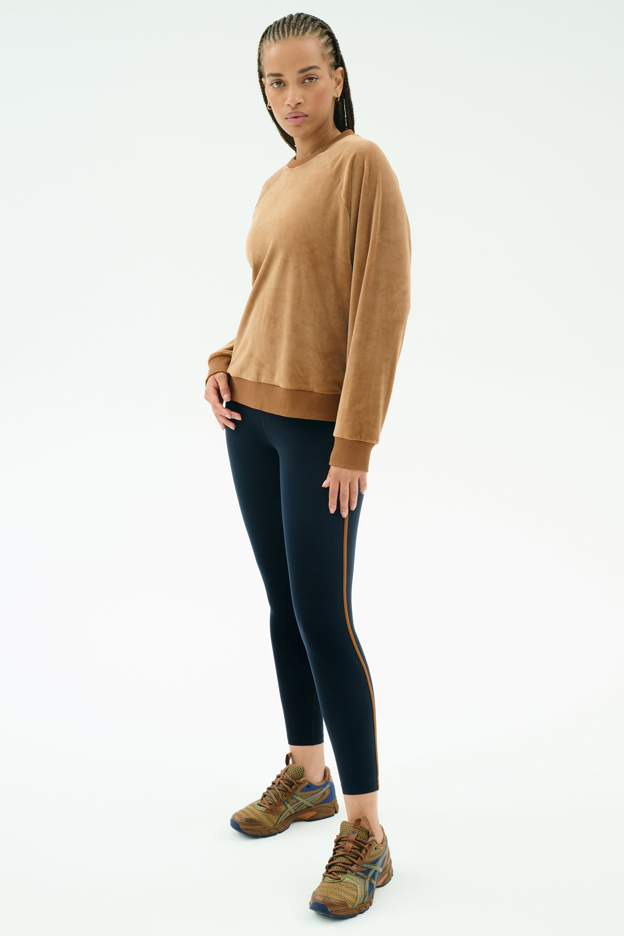 A person stands against a plain background wearing the SPLITS59 Joy Velour Crewneck in Honey, paired with black side-stripe leggings and brown athletic shoes.