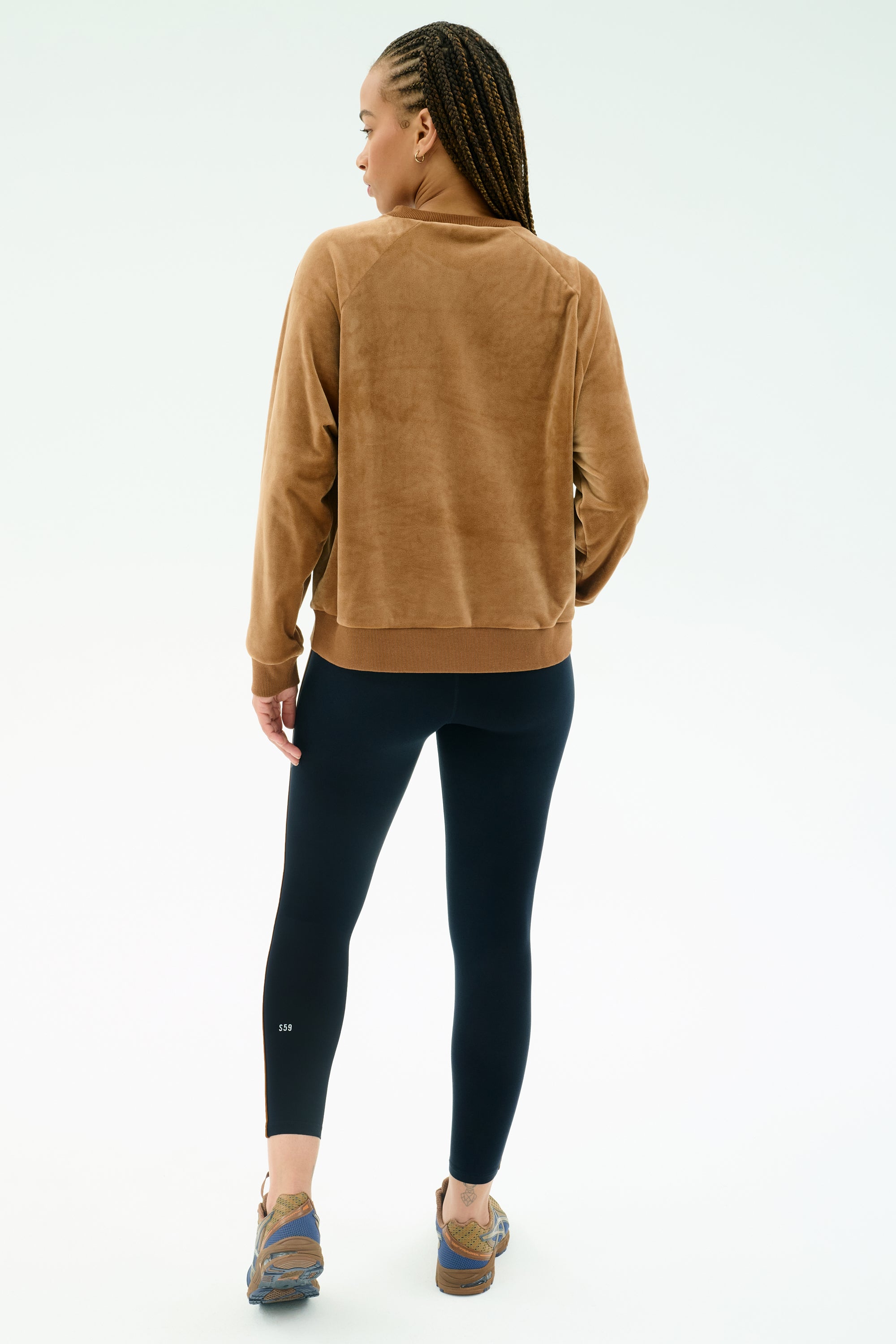 A person with braids wears the SPLITS59 Joy Velour Crewneck in Honey and black stretch leggings, standing with their back to a light background.