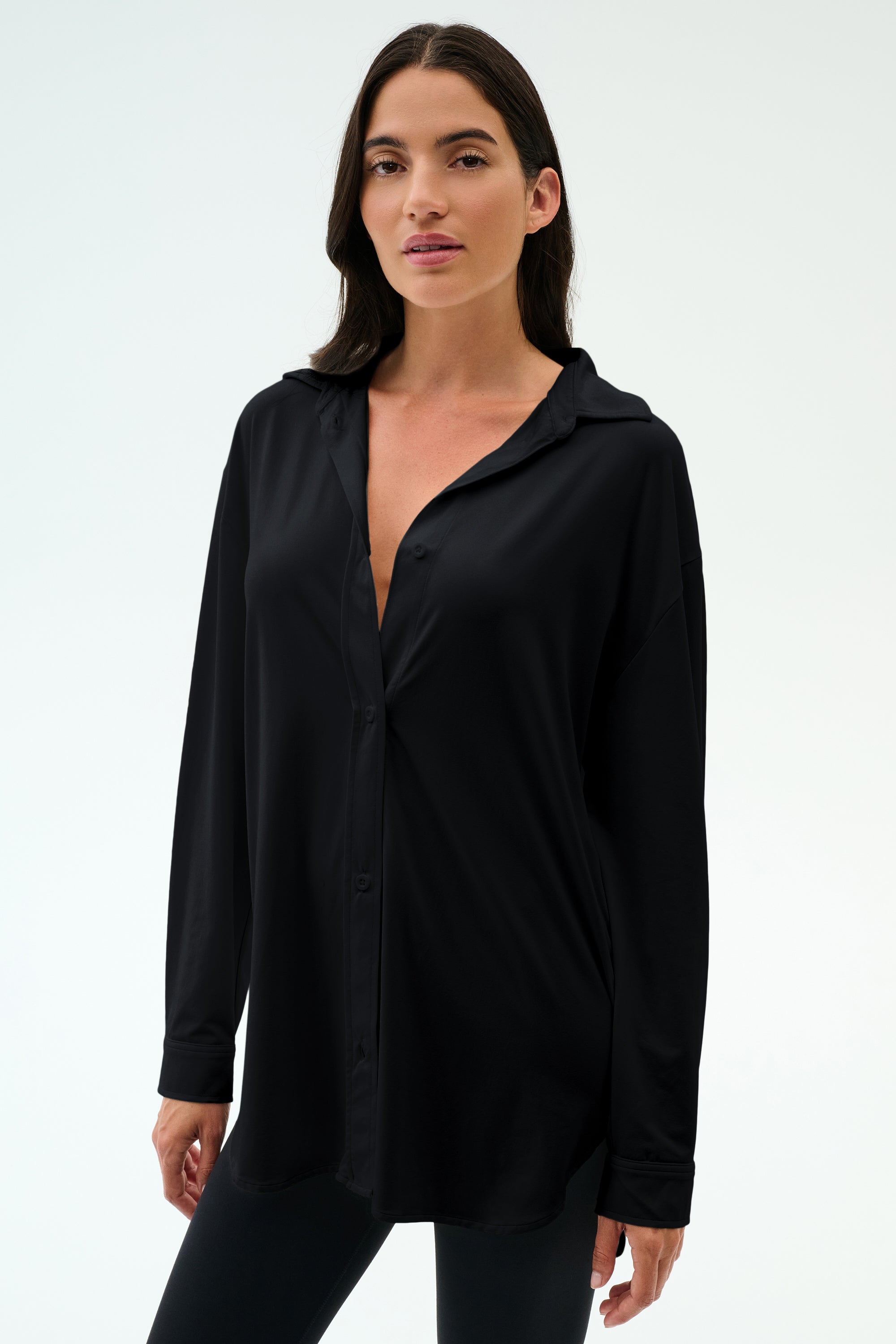 A woman with long dark hair wears the SPLITS59 Kate Airweight Lite Button Down in black with matching pants, standing against a plain light background—a versatile layering piece.