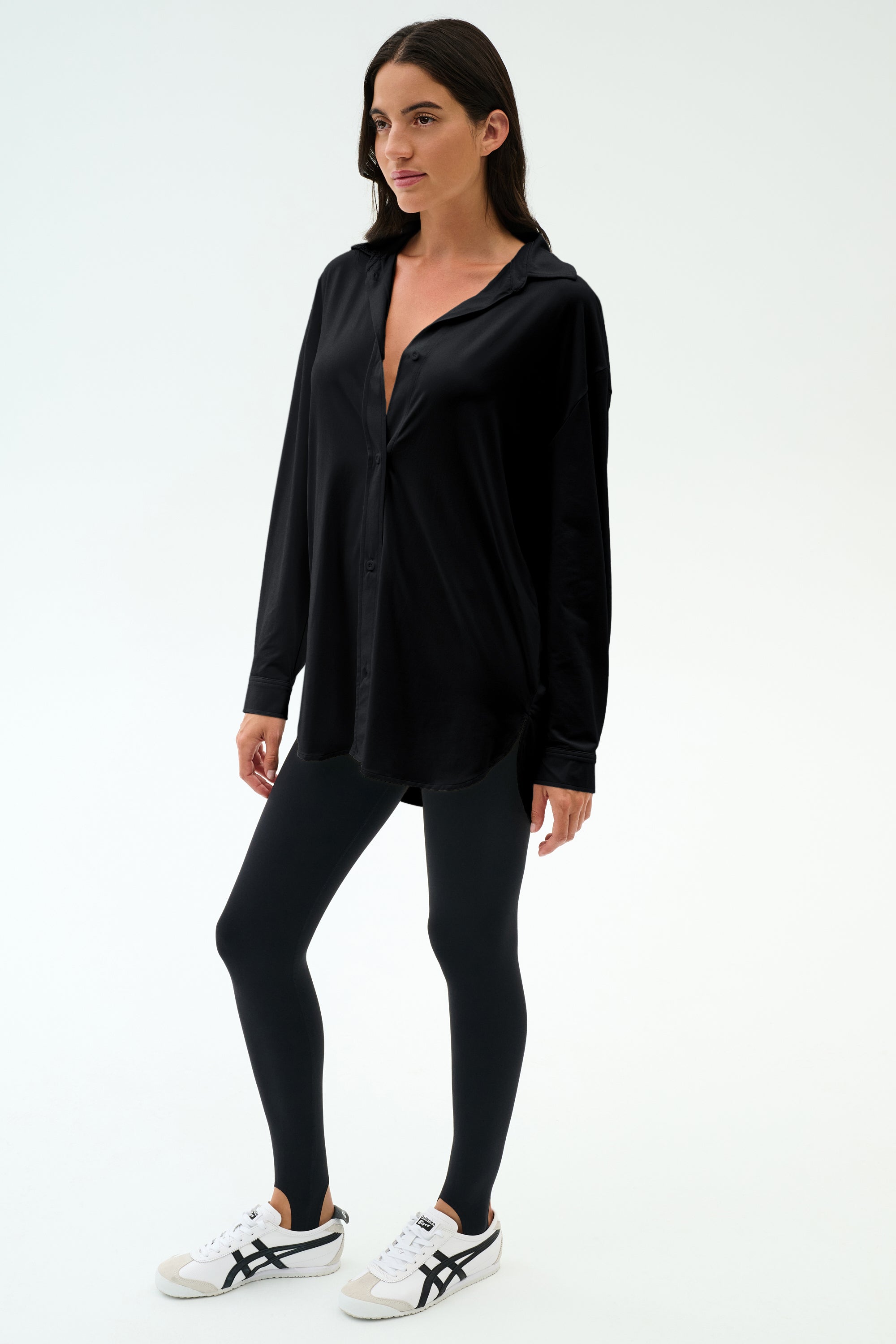 A woman stands against a plain background wearing the SPLITS59 Kate Airweight Lite Button Down in black, paired with black leggings and white sneakers with black accents—a versatile layering piece for any outfit.