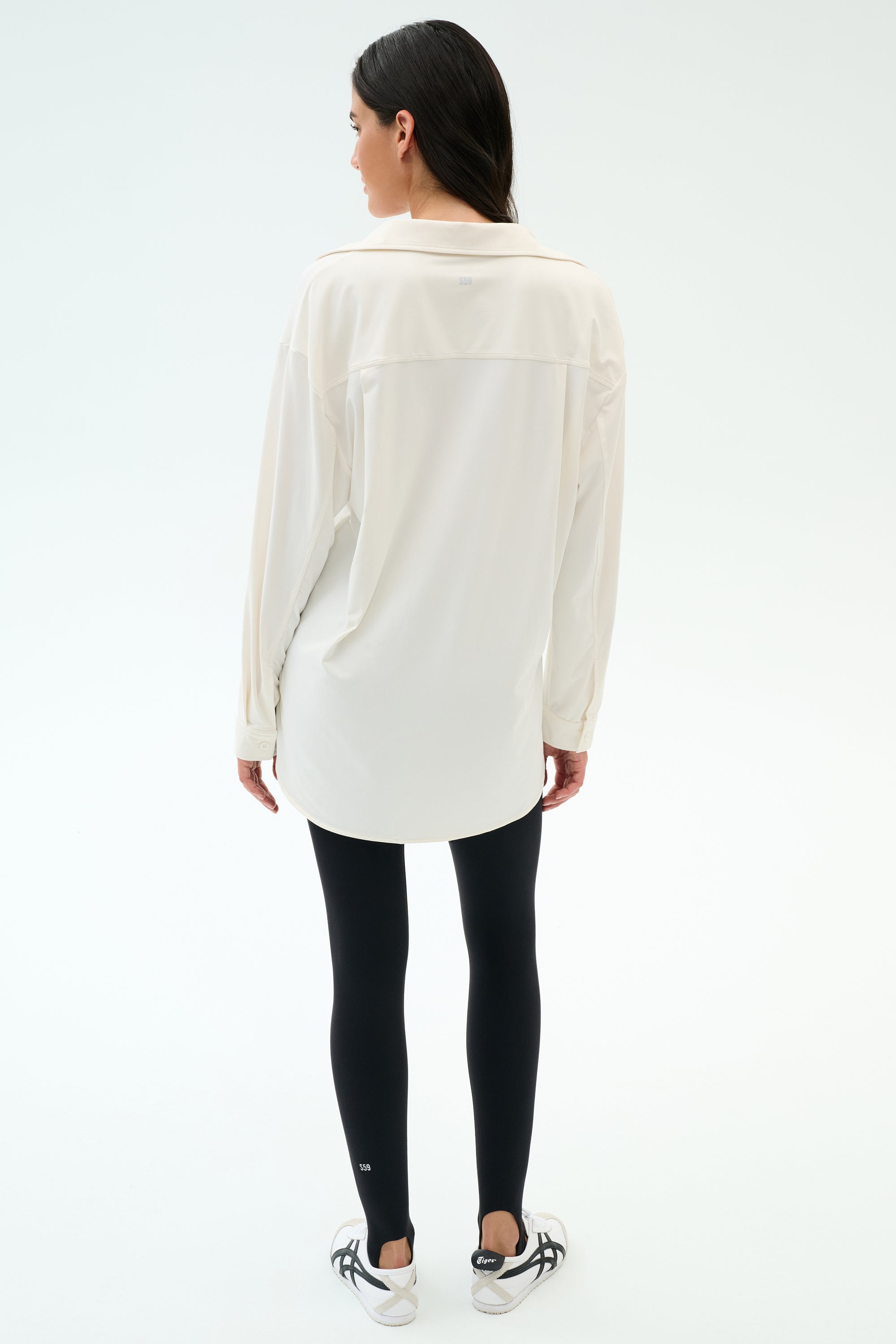 A woman faces away against a plain light background, wearing the SPLITS59 Kate Airweight Lite Button Down in white, paired with black leggings and white sneakers.