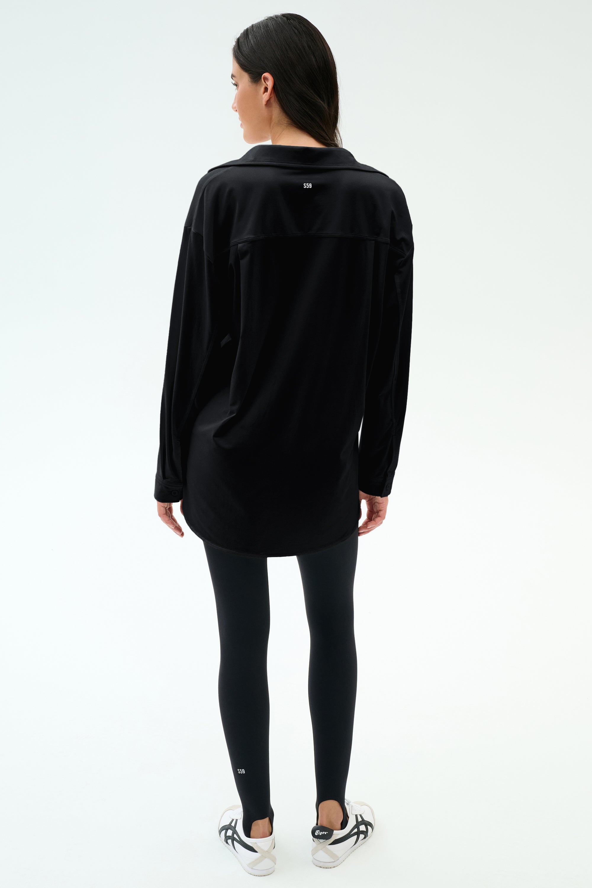 A woman faces away, wearing the SPLITS59 Kate Airweight Lite Button Down with black leggings and white sneakers against a plain, light background.