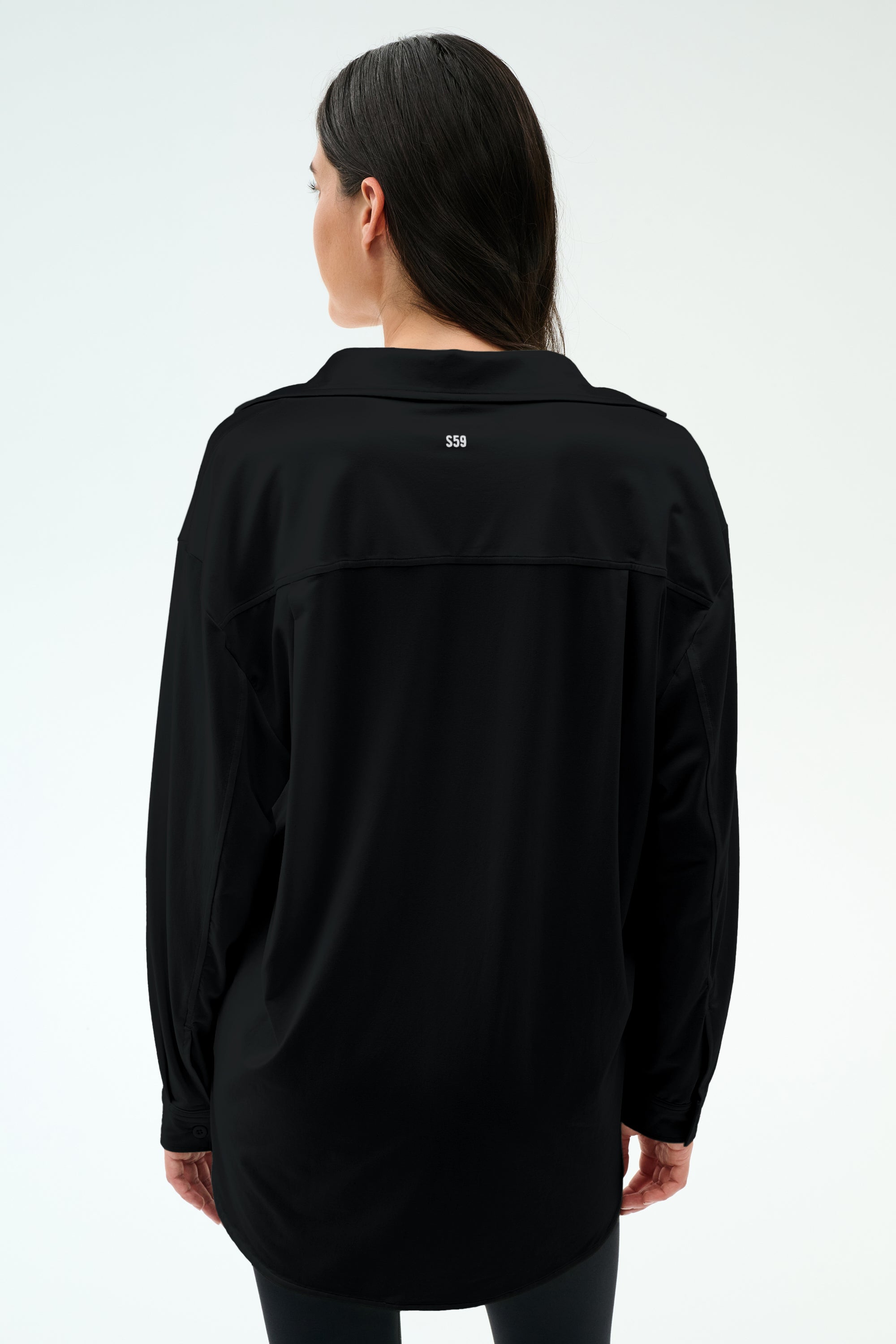 A woman with long dark hair stands with her back to the camera against a plain light background, wearing the SPLITS59 Kate Airweight Lite Button Down in black and matching pants—a breezy layering piece.