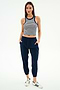 A woman faces forward in a sleeveless striped crop top, SPLITS59 Megan Woven Nylon Jogger w/ Piping in Indigo/Creme, and white Adidas sneakers with blue stripes, posed against a plain white background.