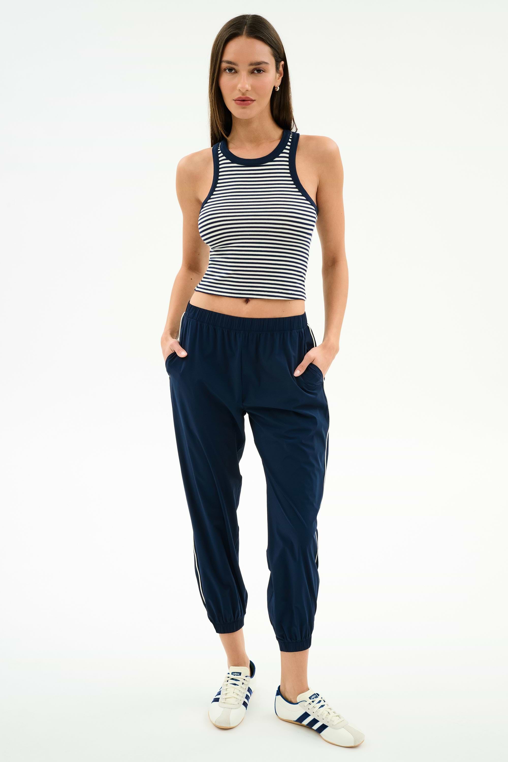 A woman faces forward in a sleeveless striped crop top, SPLITS59 Megan Woven Nylon Jogger w/ Piping in Indigo/Creme, and white Adidas sneakers with blue stripes, posed against a plain white background.