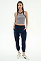 A woman faces forward in a sleeveless striped crop top, SPLITS59 Megan Woven Nylon Jogger w/ Piping in Indigo/Creme, and white Adidas sneakers with blue stripes, posed against a plain white background.