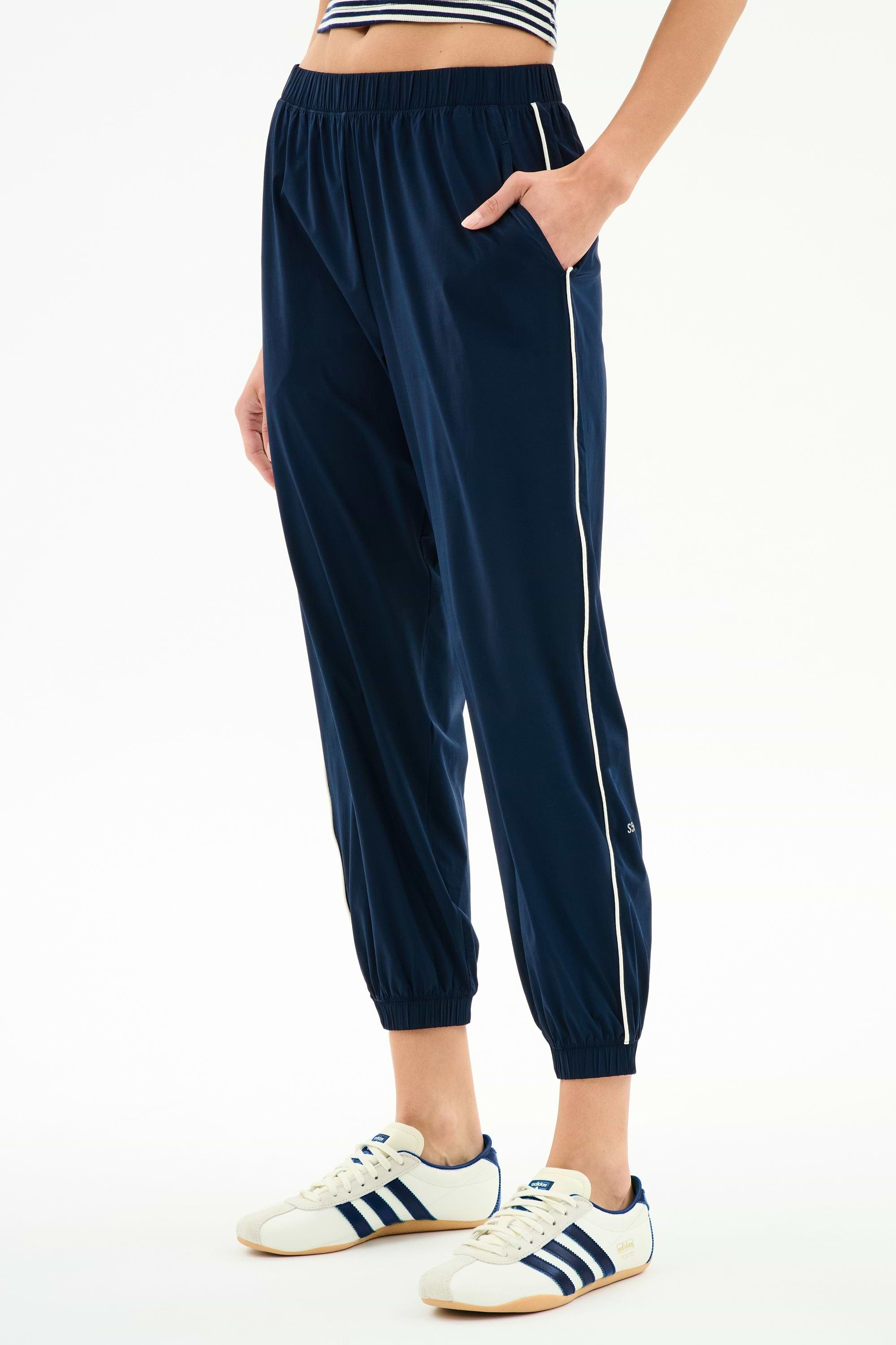 Someone wearing the SPLITS59 Megan Woven Nylon Jogger w/ Piping in Indigo/Creme, styled with a striped top and white Adidas sneakers with navy stripes, standing against a white background.