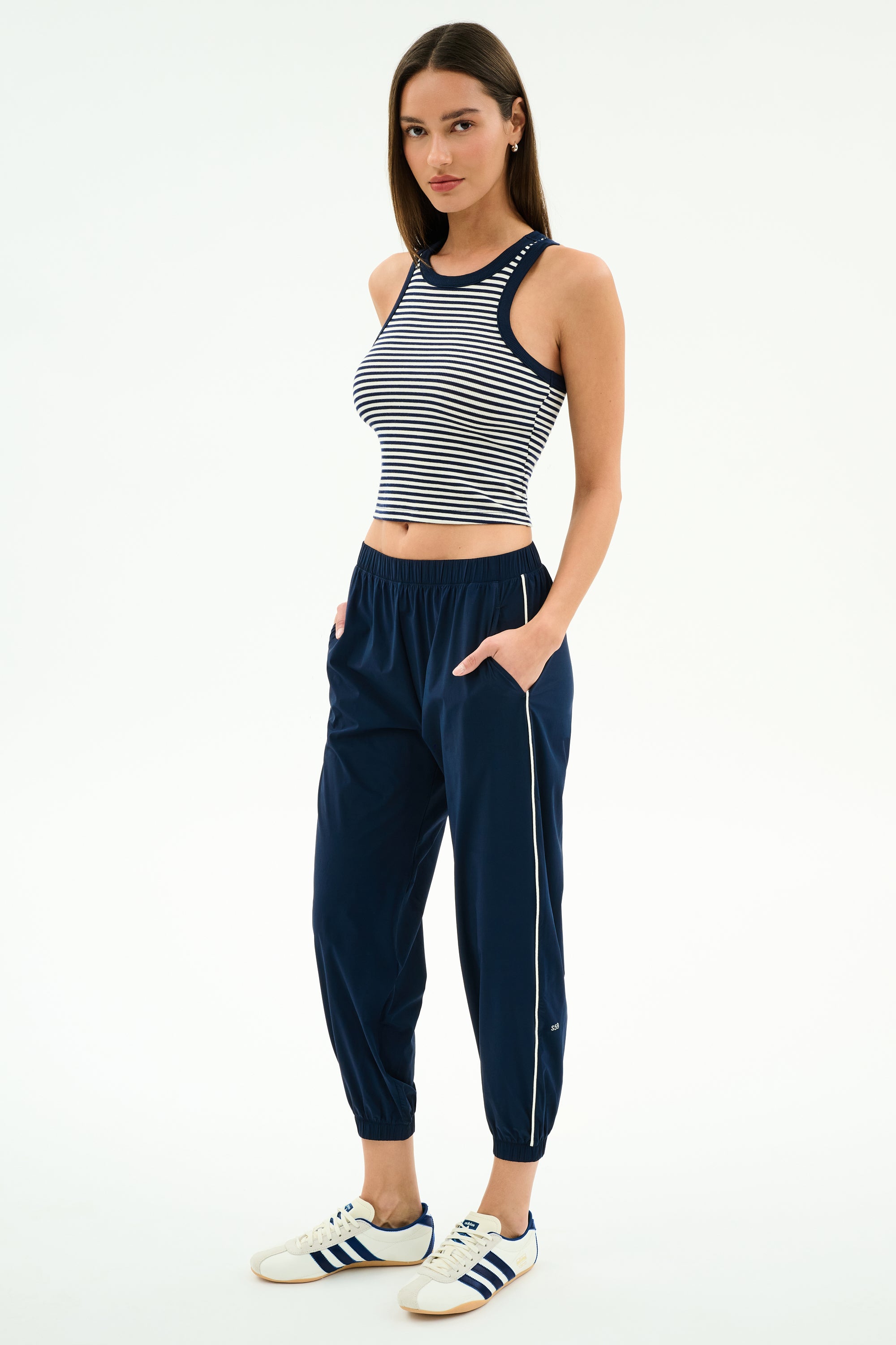 A woman stands with hands in pockets, wearing the SPLITS59 Kiki Rib Crop Tank in White/Indigo Stripe, navy jogger pants, and white sneakers with blue stripes against a plain background.