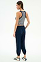A woman stands slightly turned away, wearing the SPLITS59 Megan Woven Nylon Jogger w/ Piping in Indigo/Creme, a striped sleeveless crop top, and white sneakers with navy stripes against a plain background.