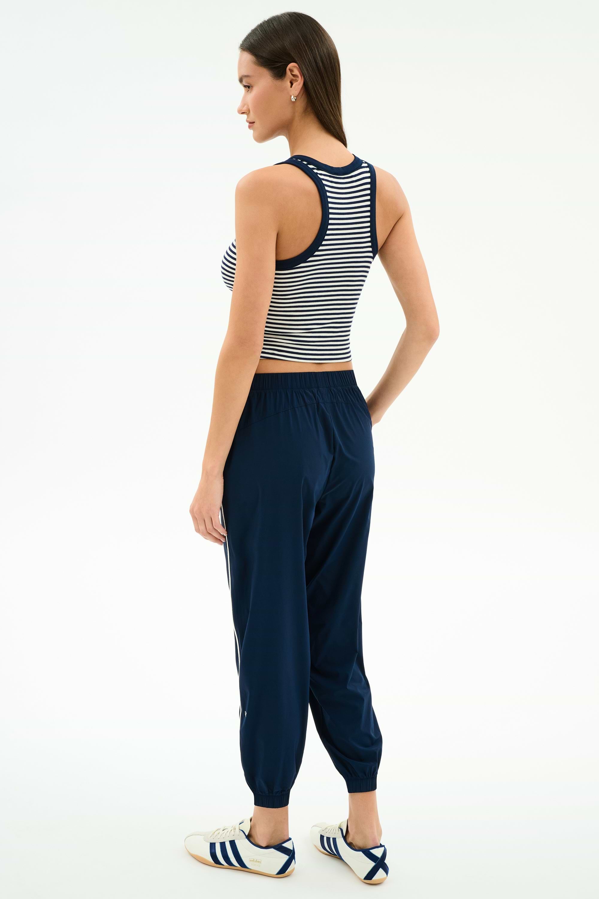 A woman stands slightly turned away, wearing the SPLITS59 Megan Woven Nylon Jogger w/ Piping in Indigo/Creme, a striped sleeveless crop top, and white sneakers with navy stripes against a plain background.