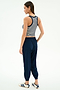 A woman stands slightly turned away, wearing the SPLITS59 Megan Woven Nylon Jogger w/ Piping in Indigo/Creme, a striped sleeveless crop top, and white sneakers with navy stripes against a plain background.
