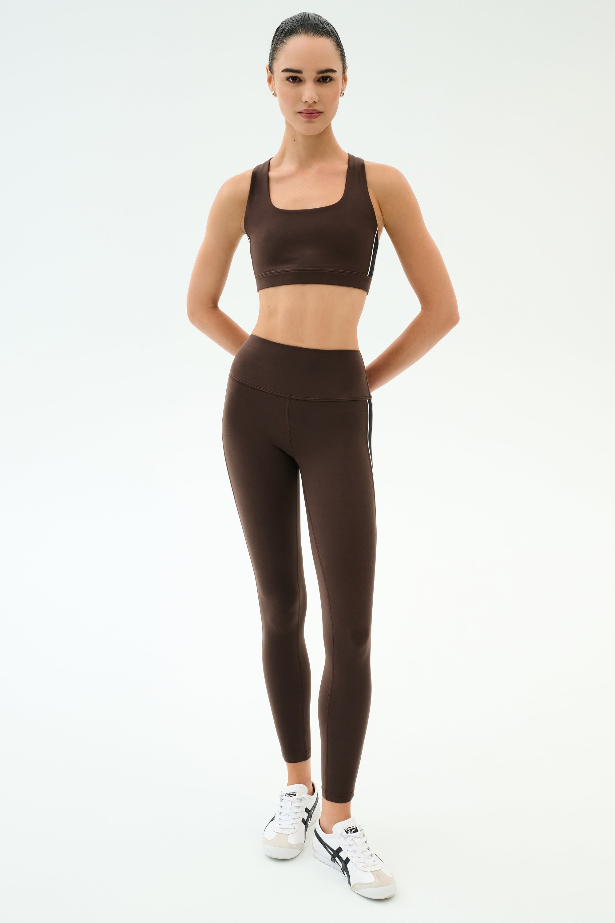 A woman stands facing forward in a SPLITS59 Margot Rigor Legging - Dark Chocolate/Black, brown sports bra, and white sneakers against a plain white background.
