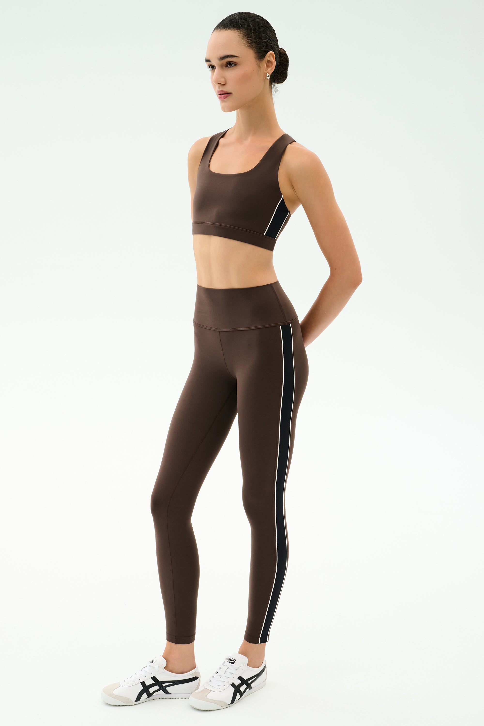 A person wears a SPLITS59 Margot Rigor Legging in Dark Chocolate/Black with side stripes, paired with a matching brown sports bra and white athletic shoes, standing against a plain white background.