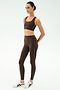 A person wears a SPLITS59 Margot Rigor Legging in Dark Chocolate/Black with side stripes, paired with a matching brown sports bra and white athletic shoes, standing against a plain white background.