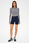 A woman stands against a white background wearing the SPLITS59 Louise Rib Long Sleeve Crop in White/Indigo Stripe, paired with navy blue high-waisted shorts and navy running shoes.