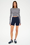 A woman stands against a white background wearing the SPLITS59 Louise Rib Long Sleeve Crop in White/Indigo Stripe, paired with navy blue high-waisted shorts and navy running shoes.