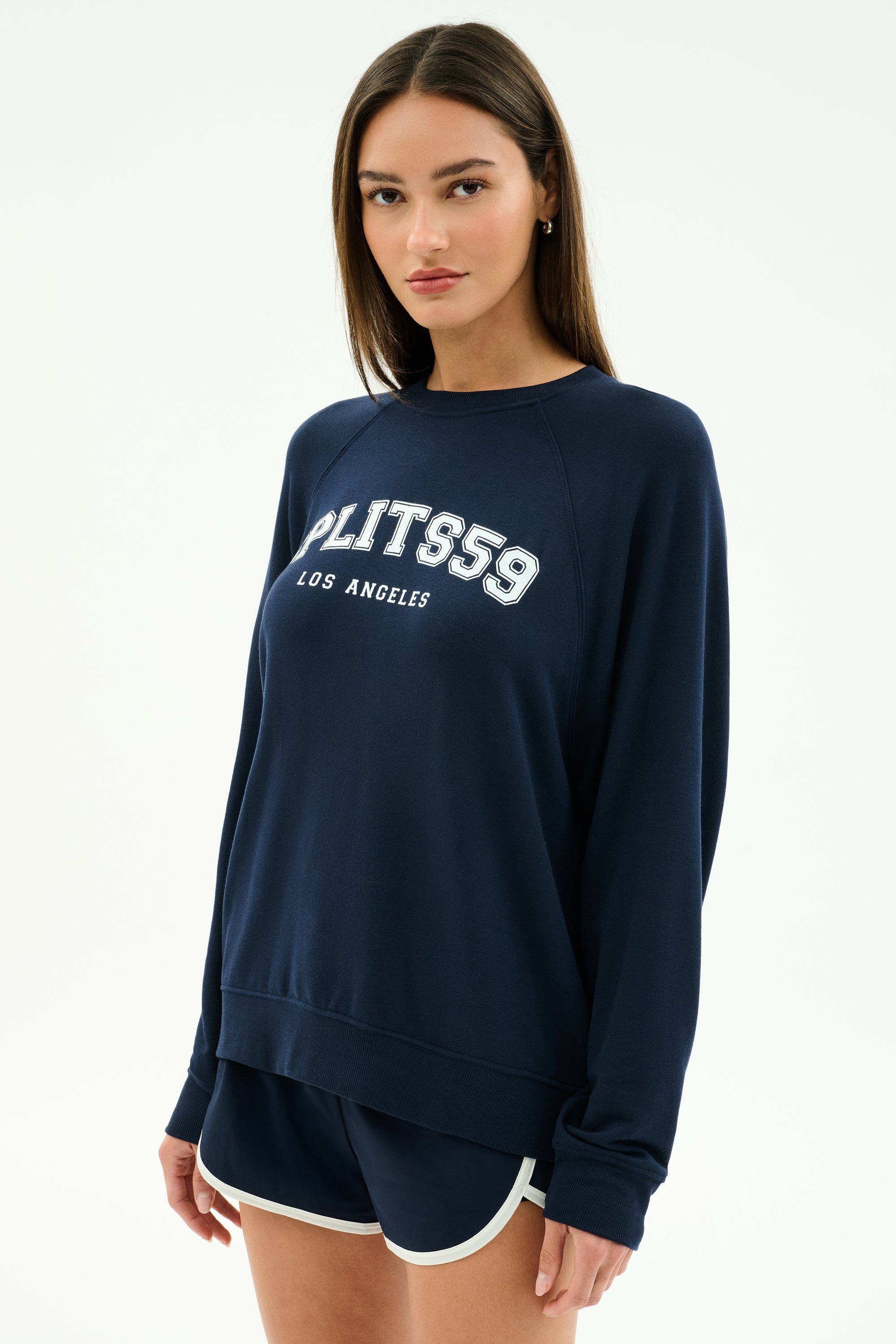 A woman wears the SPLITS59 Andie Oversized Fleece Varsity Graphic Sweatshirt in indigo and navy athletic shorts with white trim, both made in Los Angeles, standing against a plain background.