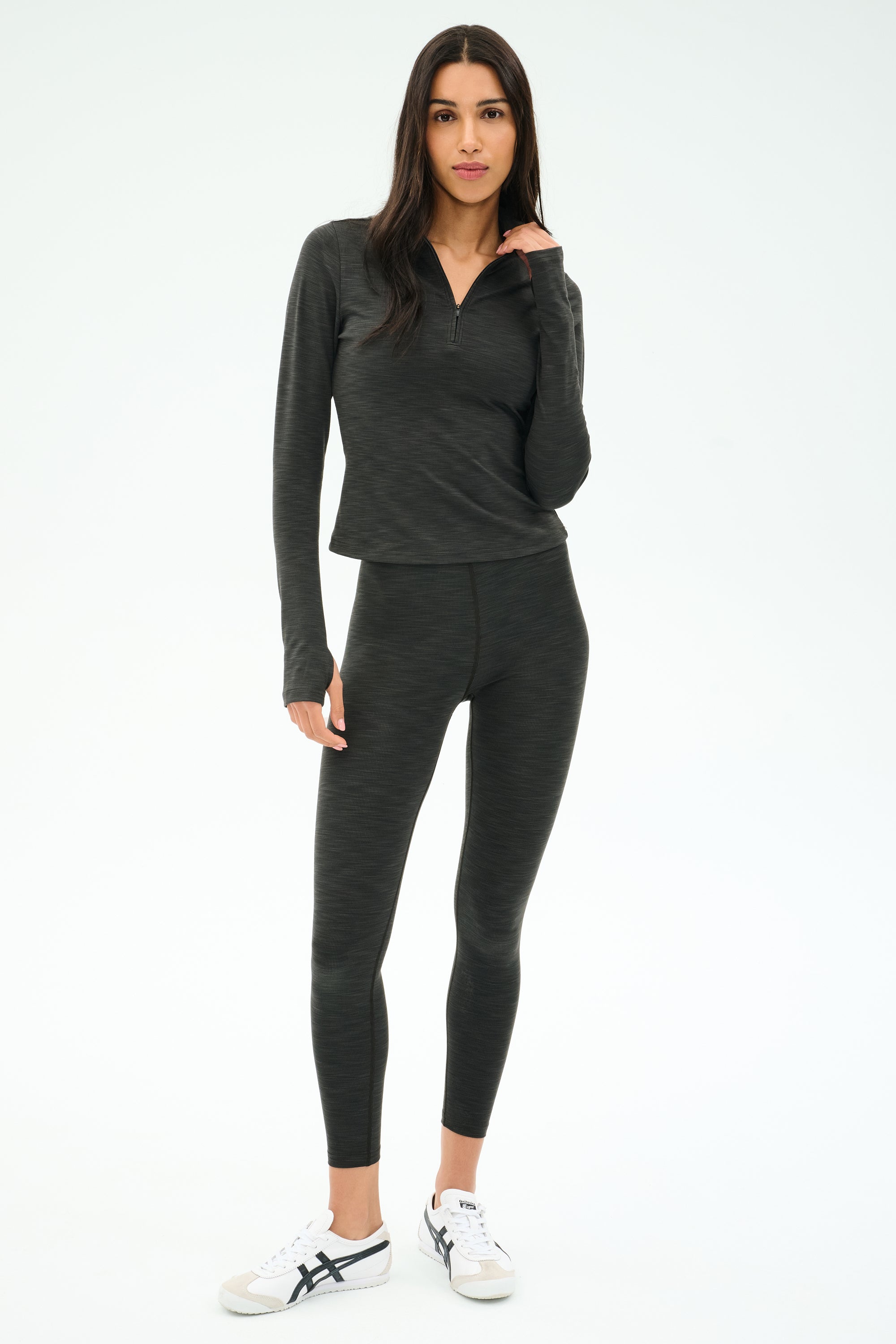 A woman stands in a studio wearing the SPLITS59 Stevie Active Rib Half Zip Waist Length top in Heather Graphite, paired with matching leggings and white sneakers, with a neutral expression.