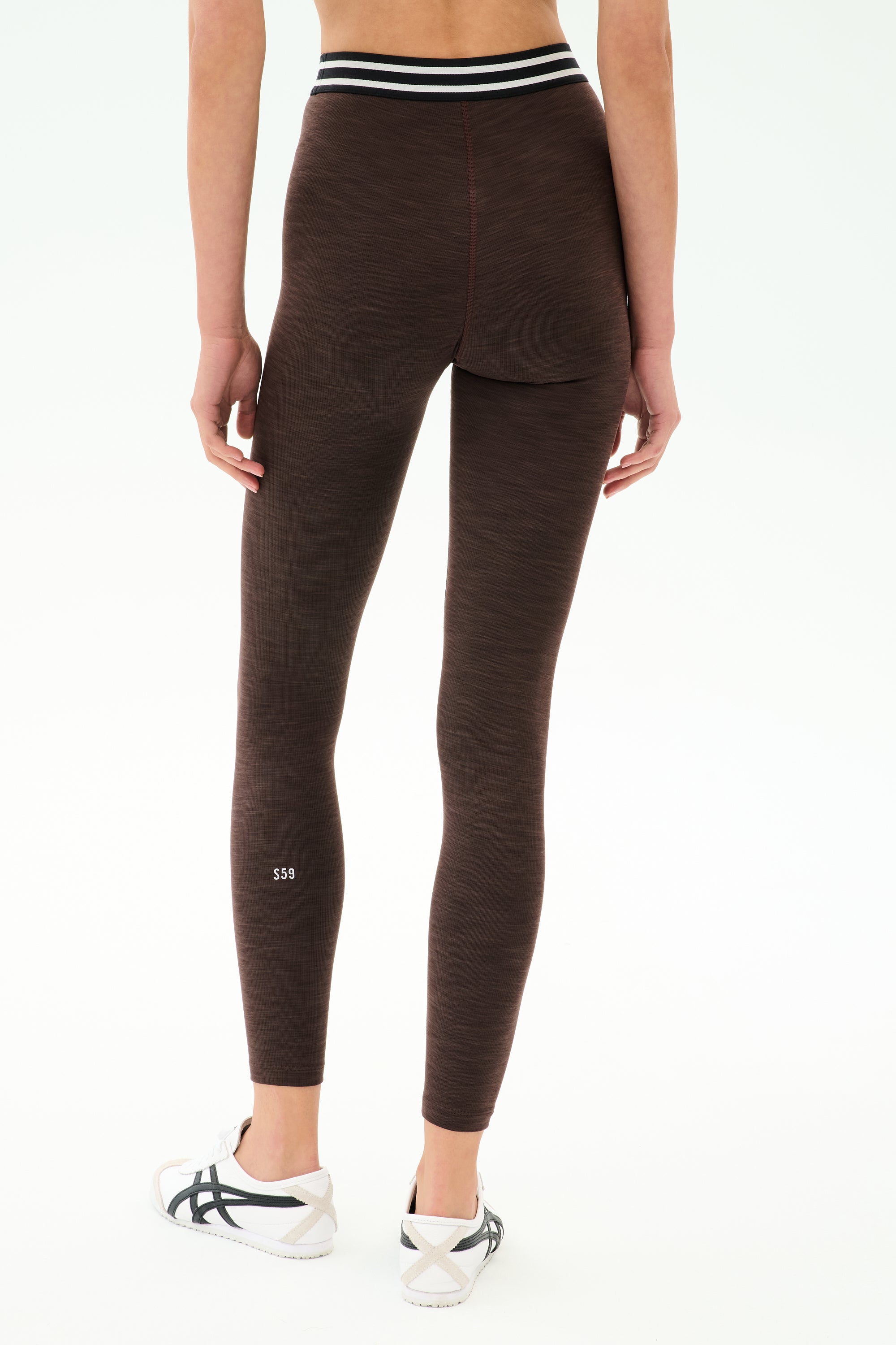 A person wears SPLITS59's Bailey High Waist Active Rib 7/8 leggings in Heather Dark Chocolate, featuring a black and white striped waistband and white sneakers, shown from the back—ideal for gym workouts.