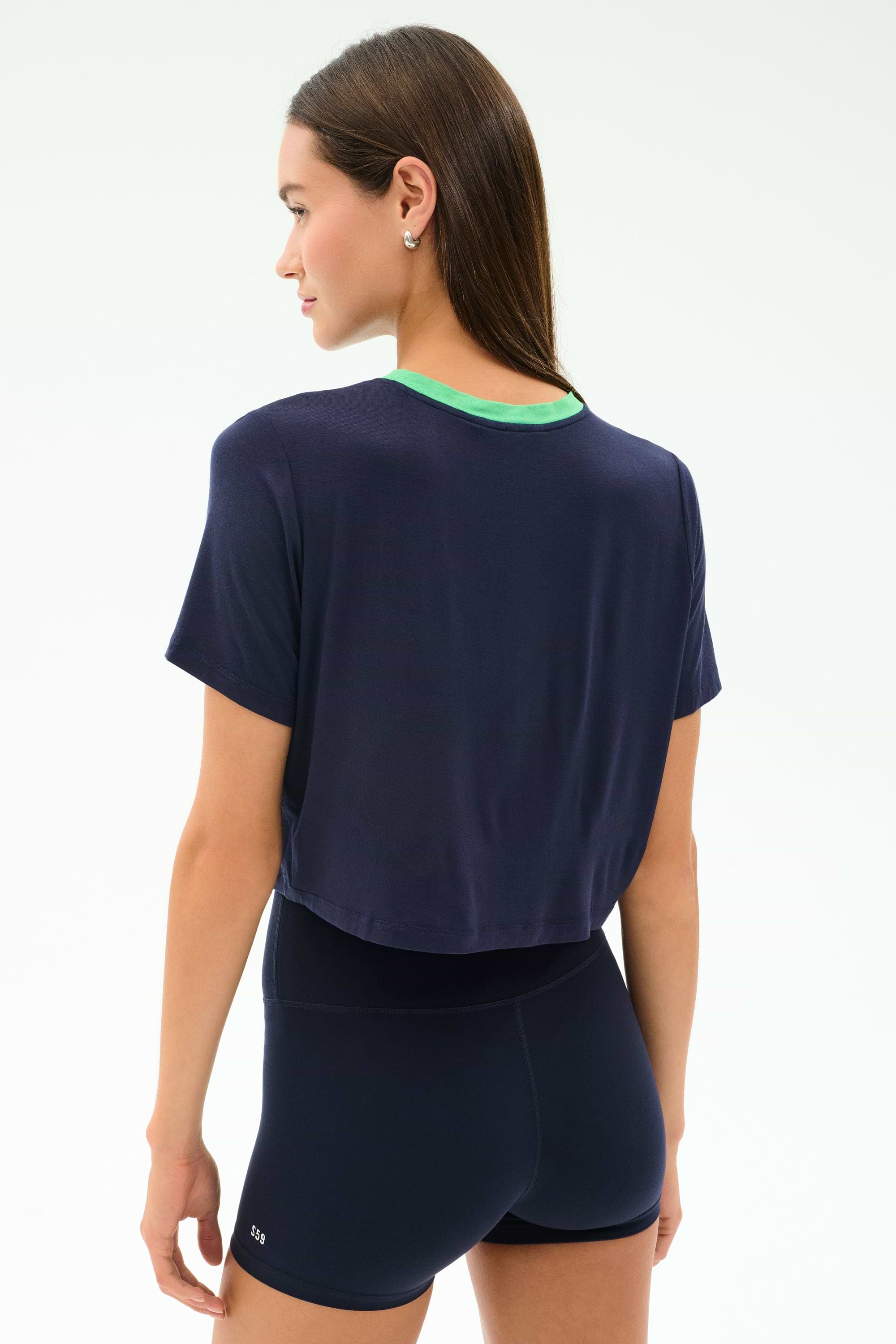 A woman faces away, wearing the SPLITS59 Djuna Crop Ringer in Indigo/Spearmint—a navy blue cropped T-shirt paired with a green top and navy blue shorts.