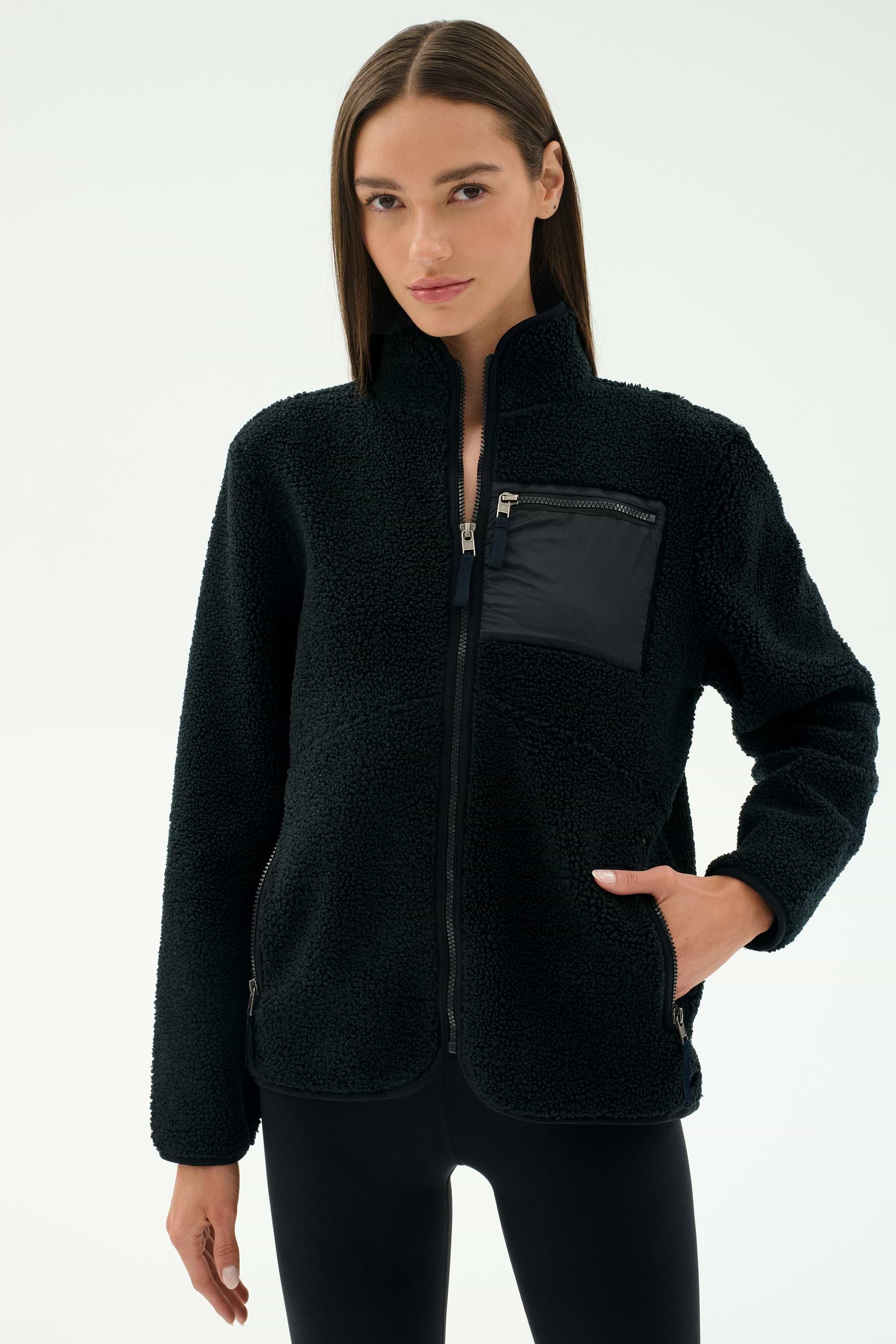 A woman stands against a plain light background wearing the SPLITS59 Harris Sherpa Jacket in Black, featuring a full zip and zippered chest pocket, paired with matching black pants—ideal for cool weather workouts.