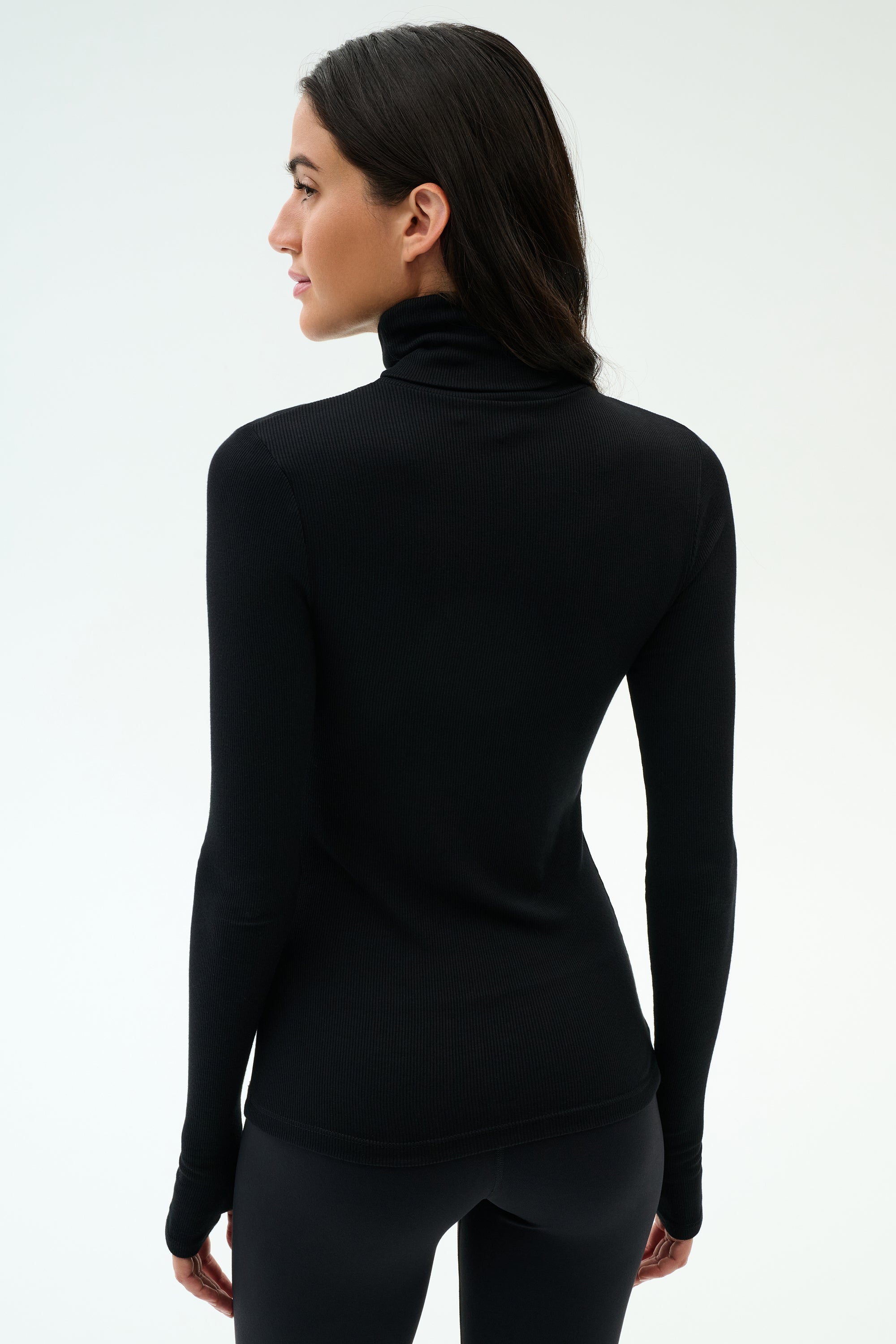 Woman stands facing away, wearing the SPLITS59 Lennon Rib Turtleneck in Black/White—sleek and fitted, ideal for yoga or running—paired with black pants against a plain light background.