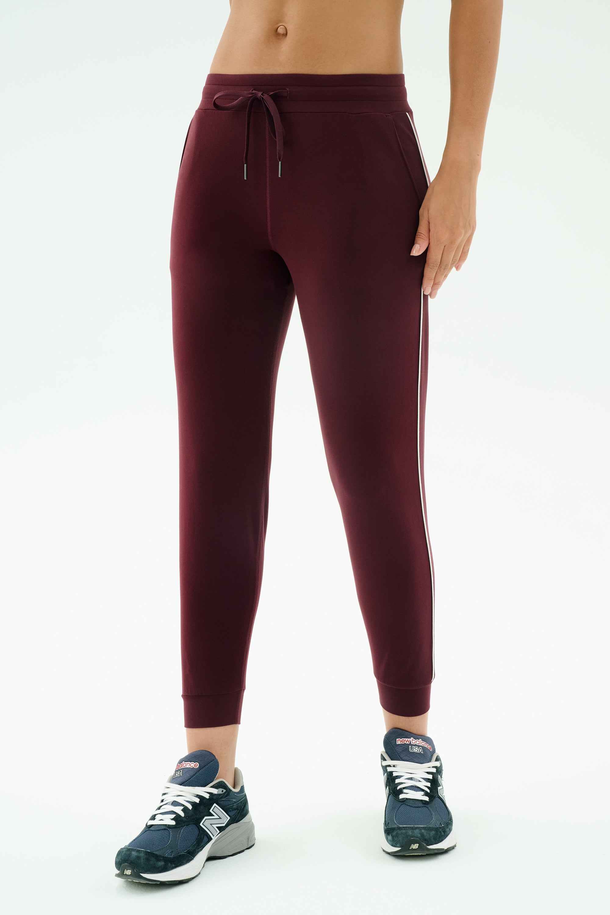 A person wears SPLITS59’s Airweight Jogger 7/8 with Piping in Dark Cherry/Indigo and dark blue sneakers, standing against a plain white background.
