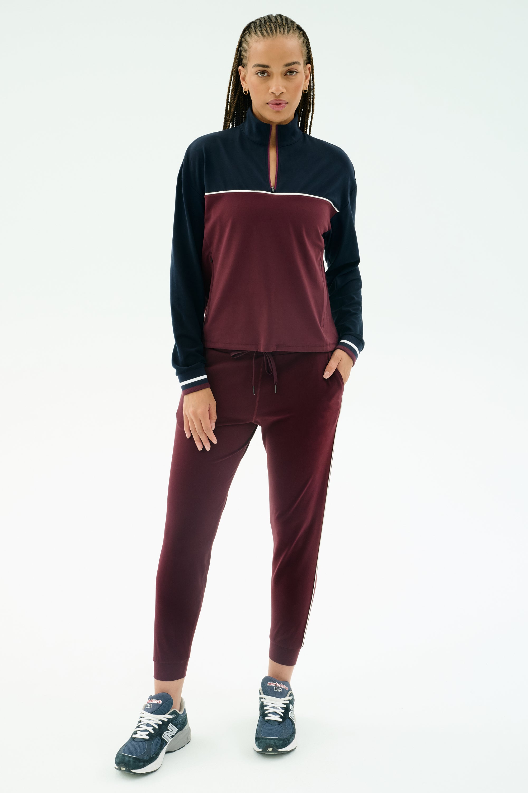 A person faces forward against a white background wearing the SPLITS59 Millie Airweight Half Zip in Dark Cherry/Indigo with matching leggings and navy sneakers.